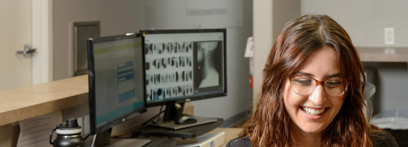 a clinician sitting and smiling in front of a computer monitor with an xray on it